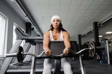 Genç bir kadın halterli ve kavisli bir halterle kolları için egzersiz yapıyor, bankta oturuyor. Spor salonundaki kadın beyaz takım elbise giyiyor ve spor yapıyor..