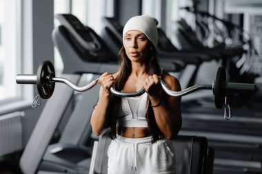 Genç bir kadın spor salonunda duran kavisli bir halterle pazı egzersizleri yapıyor. Spor salonundaki kadın beyaz takım elbise giyiyor ve spor yapıyor..