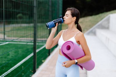 Sporcu kadın dışarıda egzersiz yaptıktan sonra su şişesinden su içiyor, elinde pembe bir yoga minderi tutuyor.