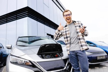 Genç bir adam, araba şarj olurken elektrikli arabasına yaslanırken cep telefonu kullanır..