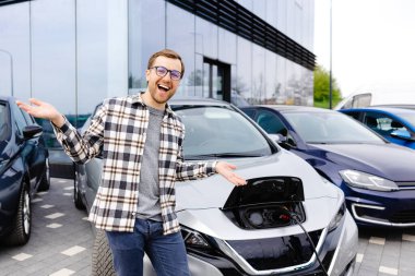 Genç bir adam yeni elektrikli arabasının yanında duruyor ve araba şarj olurken dinleniyor..