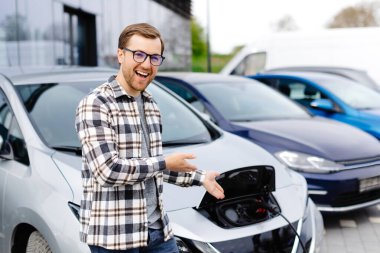 Genç bir adam yeni elektrikli arabasının yanında duruyor ve araba şarj olurken dinleniyor..