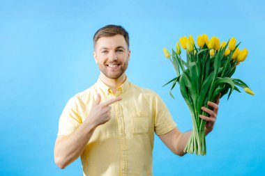 Bir adam elinde büyük bir demet parlak sarı çiçek tutuyor. Çiçekler canlı ve erkek kıyafetlerine karşı kontrastlı..