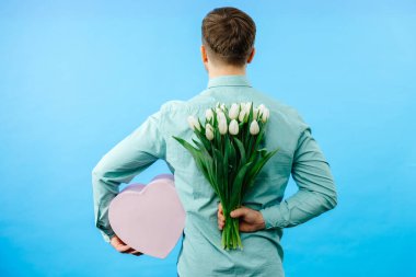 Elinde lale buketi ve hediye kutusu olan bir adam mavi arka planda izole edilmiş kameraya bakıyor. Kutlama konsepti.