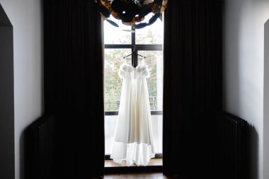 Pencerenin önündeki askıda asılı beyaz gelinlik siyah perdeli ve radyatörlü karanlık bir odada gelinin düğün gününde giymesi için hazır.