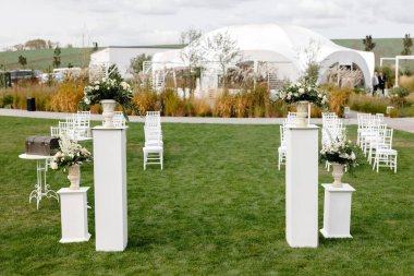 Elegant outdoor wedding ceremony setup featuring white columns with floral arrangements, white chairs on lush green lawn, and reception tent clipart