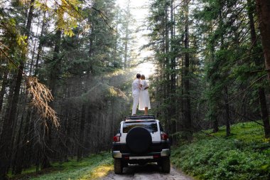 Couple standing on the roof of their suv embracing and enjoying the view in a forest during a road trip clipart