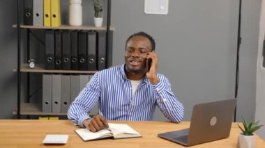 Afrikalı yönetici ofis masasında dizüstü bilgisayarın önünde oturuyor ve keyifli bir telefon görüşmesi yapıyor..