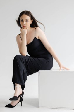 Beyaz bir kübün üzerinde oturan ve poz veren zarif siyah elbiseler giyen, yüksek topuklu genç beyaz bir kadının stüdyo fotoğrafı.