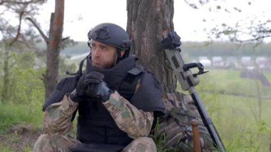 Askeri üniformalı ve kurşun geçirmez yelek giyen bir adam ormanda bir metal dedektörünün ve askeri bir sırt çantasının yanında oturuyor. Bir adam bölgeyi mayınlardan arındırmak için duraksadı.