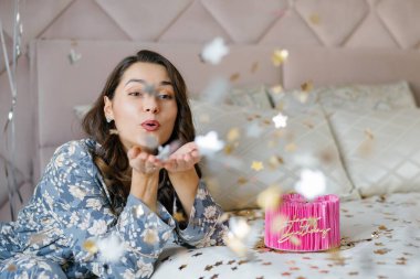 Güzel genç bir kadın doğum günü pastasına altın konfeti üflüyor, pijamalarıyla yatağa uzanıyor, doğum gününü evde kutluyor.