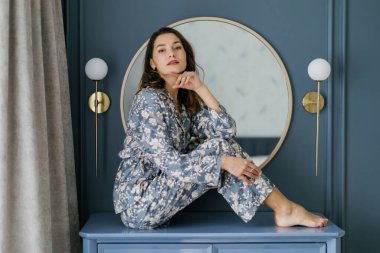 Young woman wearing pajamas is sitting on a piece of furniture in her bedroom, with a round mirror and wall lamps in the background clipart