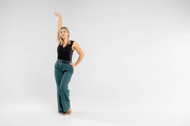 Stüdyoda, elleri beyaz bir arka planda, zarif kıyafetler giymiş, kendinden emin ve profesyonel görünümlü genç sarışın bir kadın görülüyor.