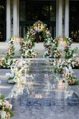 Çiçekler, avizeler ve yansıtıcı bir yüzey ile süslenmiş şık bir açık hava düğün koridoru lüks bir ortam yaratıyor.
