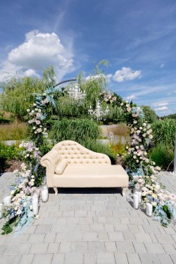 Çiçekli kemerin altında bej bir şezlonga sahip lüks bir düğün hazırlığı, romantik bir açık hava ortamı yaratıyor.