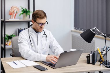 Doctor man sitting at the desk at his working place and working on laptop. Perfect medical service in clinic. Happy future of medicine and healthcare.
