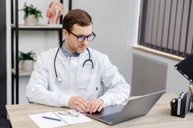 Young attractive otolaryngologist doctor sitting at his workplace in the office and holding a nose mirror.