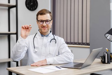 Genç, çekici bir doktor, ofisteki işyerinde oturuyor ve elinde bir kulak hortumu tutuyor..