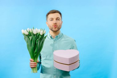 Elinde lale buketi ve hediye kutusu olan bir adam mavi arka planda izole edilmiş kameraya bakıyor. Kutlama konsepti.