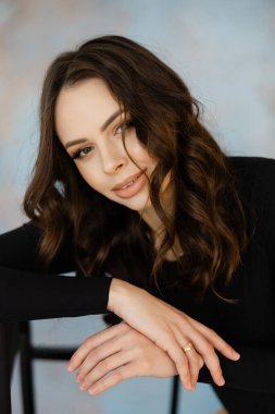 Uzun dalgalı saçları ve kendine güveni olan genç bir kadın, bir sandalyeye yaslanmış, stüdyo ortamında yumuşak, bulanık bir arka plan üzerine kurulmuş.