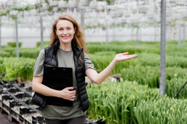 Kitapları olan genç bir bayan işçi lalelerle serada duruyor..