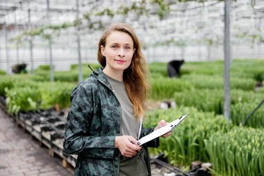 Kitapları olan genç bir bayan işçi lalelerle serada duruyor..