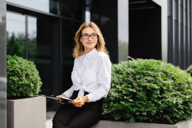 Zarif, kendine güvenen genç bir kadın, resmi olarak giyinmiş, iş merkezinin arka planında duruyor, elinde belgelerle birlikte bir klasör tutuyor, kameraya bakıyor..