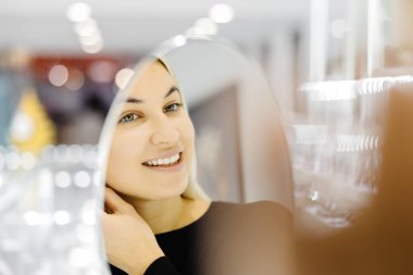 Bir kuyumcunun aynasında kendine bakarken yeni altın küpeler deneyen gülümseyen sarışın kadın.