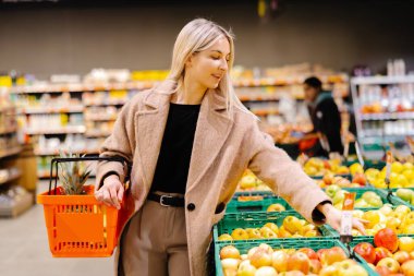 Süpermarketten alışveriş yaparken elinde elma ve alışveriş sepeti tutan gülümseyen kadın.