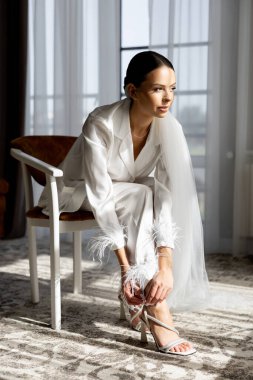 Beyaz bir takım elbise ve duvak giyip yüksek topuklu ayakkabılarını bağlayan, güneşli bir otel odasında bir sandalyede oturan, düğün gününe hazırlanan zarif gelin.