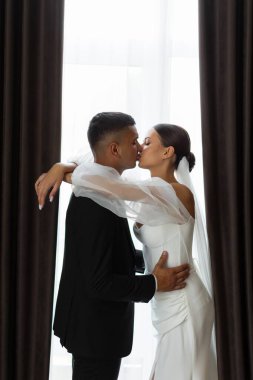 Düğün günü pencerenin yanında gelini alnından öperken romantik bir portre.