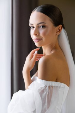 Güzel gelin peçeli beyaz gelinlik giyer pencerenin yanında gözleri kapalı poz verir.