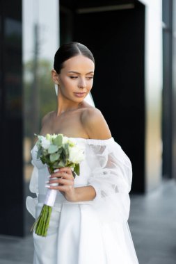 Güzel gelin, modern bir binanın yanında beyaz bir buketle poz veriyor. Özel düğün gününde neşe ve zarafet saçıyor.