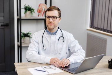 Young attractive otolaryngologist doctor sitting at his workplace in the office and holding a nose mirror.