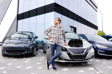 Genç bir adam yeni elektrikli arabasının yanında duruyor ve araba şarj olurken dinleniyor..