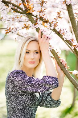 Parkta çiçek açan sakura ağacının yanında poz veren sarı saçlı çekici genç bir kadının portresi.