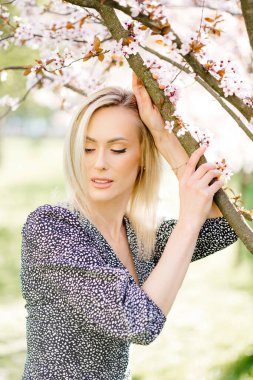 Parkta çiçek açan sakura ağacının yanında poz veren sarı saçlı çekici genç bir kadının portresi.