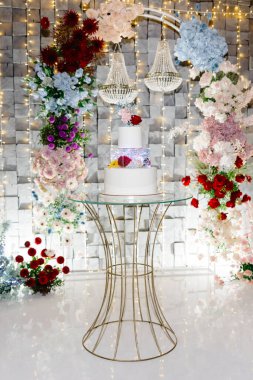 Renkli çiçekler ve zarif avizelerle çevrili, altın bir stantta sergilenen güzel beyaz düğün pastası şenlikli bir atmosfer yaratıyor.