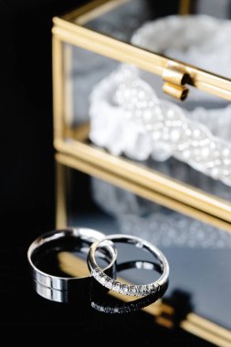 Zarif alyanslar ve jartiyer cam bir kutuda sergilenir, yanında bir buket beyaz gül, romantik ve lüks bir atmosfer yaratır.