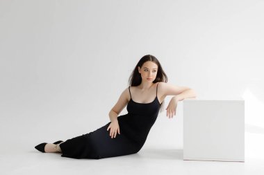 Uzun kahverengi saçlı, zarif siyah elbisesi ve beyaz arka planda duran kolyesi olan, beyaz küplere yaslanmış genç bir kadın.