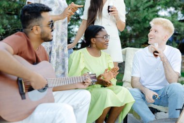 Çok ırklı bir grup insan, gitar çalan Hintli bir adam ve arkadaşları piknik sırasında yemek yiyip şarap içiyorlar..