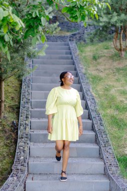 Koyu tenli bayan dışarıda gülüyor. Sarı elbiseli esmer kadın gülümsüyor ve dışarı çıkıyor..