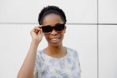 Koyu tenli bayan dışarıda gülüyor. Esmer, kıvırcık elbiseli, güneş gözlüklü kadın gülümseyip dışarı çıkıyor..