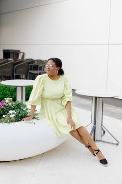 Koyu tenli bayan dışarıda gülüyor. Sarı elbiseli esmer kadın gülümsüyor ve dışarı çıkıyor..