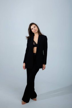 Siyah takım elbiseli, düğmesiz ceketli ve dantelli sutyen giyen genç bir girişimci stüdyoda çıplak ayakla beyaz arka planda duruyor.