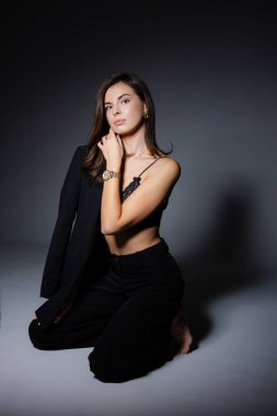 Siyah takım elbise ve dantel sutyen giymiş, gri arka planda poz veren genç bir iş kadınının stüdyo portresi.