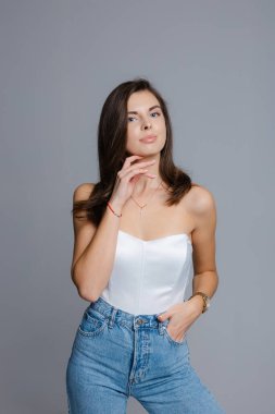 Beyaz korseli bluz ve kot pantolon giyen, elleri kalçasında poz veren ve omzunun üstünden bakan güzel bir genç kadının stüdyo fotoğrafı.