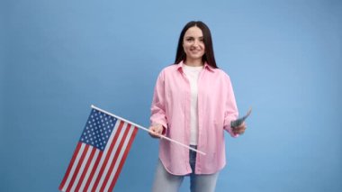 Pembe gömlekli genç esmer kadın stüdyoda mavi arka planda Amerikan bayrağı ve pasaportlu 100 dolar ile poz veriyor. Başarı kavramı, seyahat, yabancı dil bilgisi