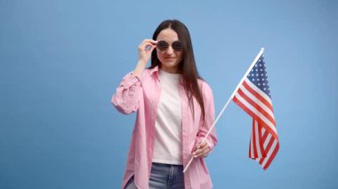 Gülümseyen mutlu genç kadın Amerikan bayrağı taşıyan günlük kıyafetler giyiyor ve mavi arka planda izole edilmiş kameraya bakıyor.
