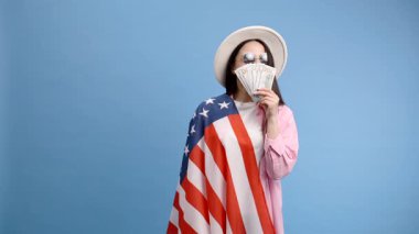 Pembe gömlekli genç esmer kadın stüdyoda mavi arka planda Amerikan bayrağı ve 100 dolar nakitle poz veriyor. Paranın kokusu.